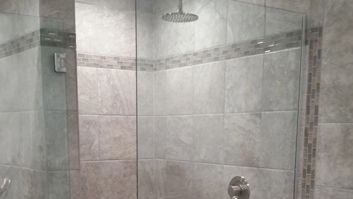 A spacious and elegantly designed shower area with large light grey Onyx tiles and a central band of decorative mosaic tiles running horizontally across the walls. 